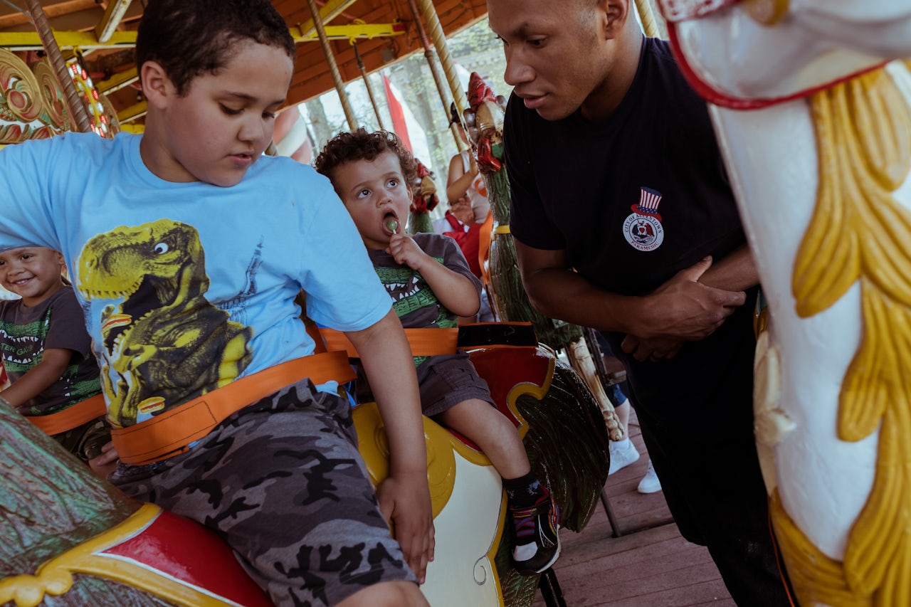 Young children donning brightly colored dinosaur shirts share a relaxing moment on the the Six Flags Columbia Carousel, accompanied by an adult.