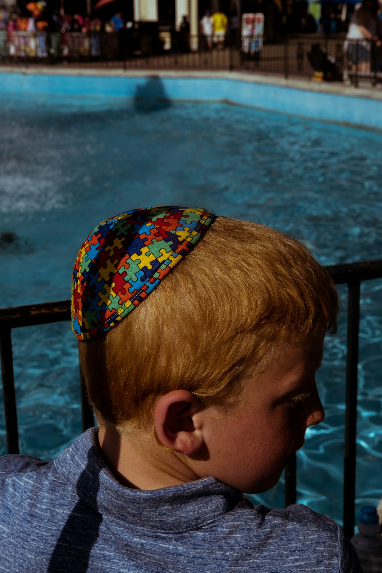 A young boy stands beside the Main Street Fountain, located near the entrance of the park, wearing a kippah bearing the multicolored jigsaw pattern that represents autism awareness.